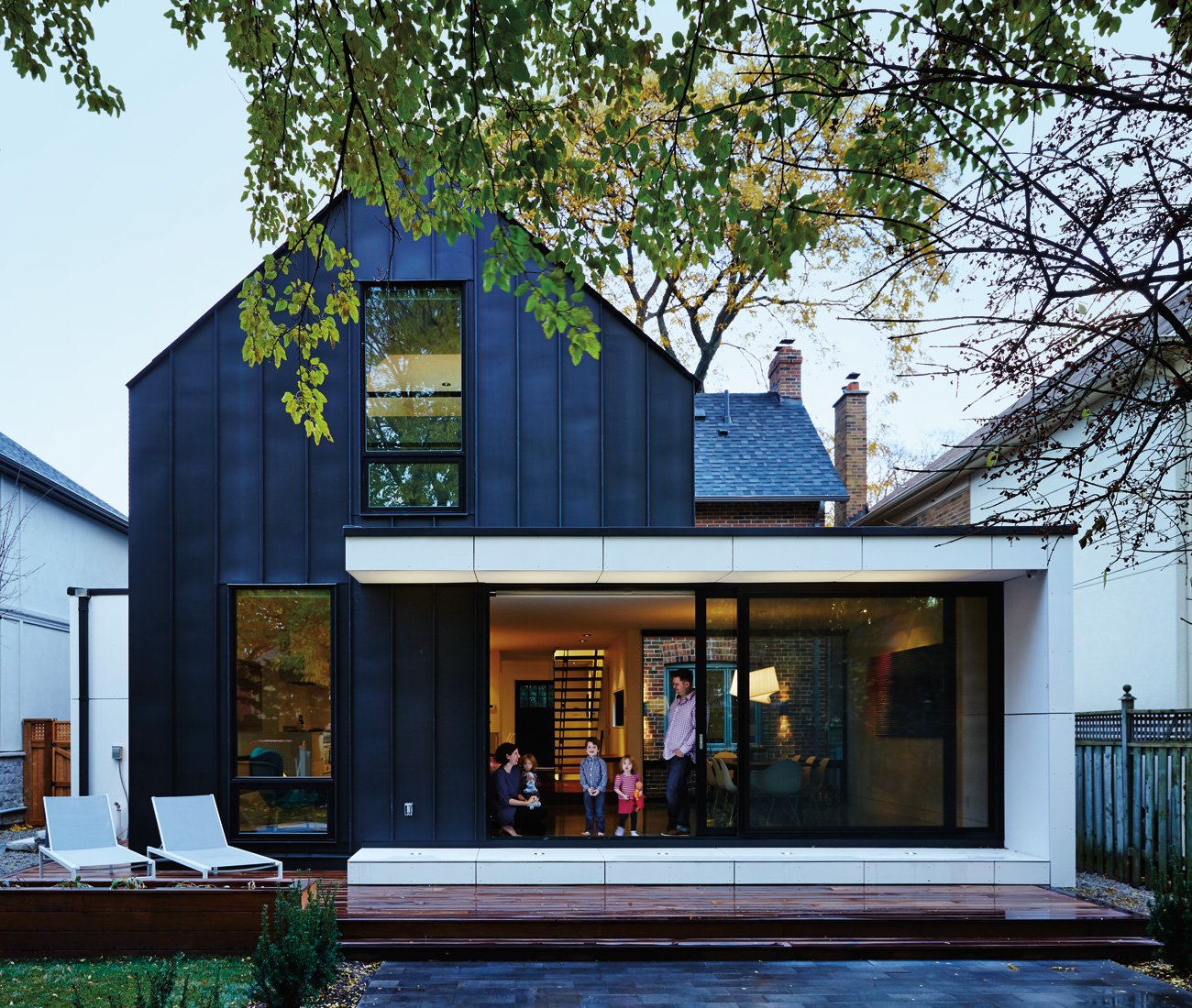 A family is standing near the sliding door facing the backyard patio. The house is cladded with zinc panels